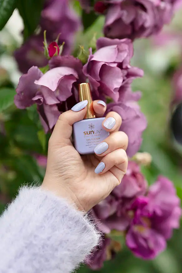A hand with lavender purple manicured nails holds a small bottle of SUNUV brand nail polish against a background of purple flowers and green leaves.